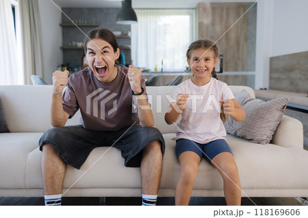Brother and sister watching TV and reacts emotionally to the events in the TV show. Teenage boy and little girl sitting on couch at home. Brother and sister watching TV and reacts emotionally to the events in the TV show. Teenage boy and little girl sitting on couch at home. 118169606