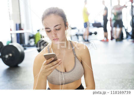 Young fitness girl checking her performance on smatphone after hard workout in gym. Young fitness girl checking her performance on smatphone after hard workout in gym. 118169739