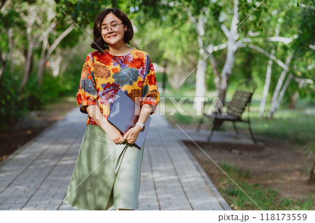 A fashionable, smiling Asian woman hugging a laptop is walking in the park. Smiling freelance worker combining work and leisure in a green urban environment 118173559