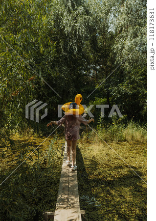 A girl is walking on a wooden bridge through a pond with a yellow duck float on her head. The photo is taken from behind, showing no face 118173631