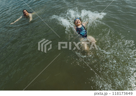 Young girl diving backwards into water with splashes under the supervision of her mother. She is enjoying a safe summer swim while her mother ensures her safety and care 118173649