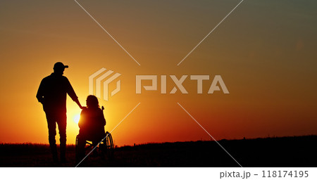 A silhouette of a person standing beside someone in a wheelchair at sunset, with a hand on their shoulder. 118174195