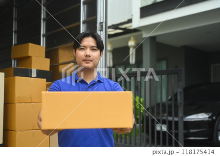 Handsome delivery man holding cardboard box standing next to a truck filled with packages 118175414