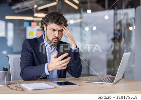 Businessman in suit experiencing frustration while looking at phone in modern office. Emotional reaction to message or email, working with laptop and tablet at desk. 118175619