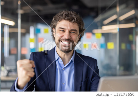 Happy confident businessman wearing a suit celebrating success in modern office. Smiling man showing joy with clenched fist gesture, conveying achievement and positive energy. 118175650