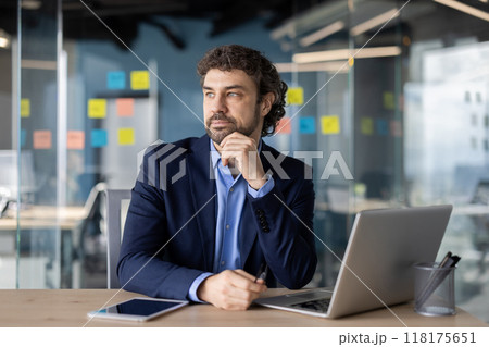 A thoughtful businessman in a modern office setting, contemplating strategy. He is surrounded by digital devices and colorful notes on a glass wall, reflecting a dynamic work atmosphere. 118175651