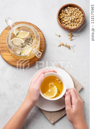 Hands holding a glass cup with chamomile herbal tea on light background with dry flowers and teapot 118175965