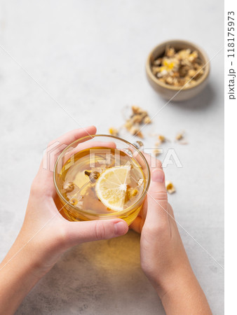 Hands holding a glass cup with chamomile herbal tea on a light background with dry flowers. Hands holding a glass cup with chamomile herbal tea on a light background with dry flowers. 118175973