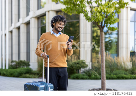 Smiling man checks phone while pulling suitcase outside modern building. Represents business travel, technology use, and professional lifestyle in casual attire, connecting work and leisure 118176412