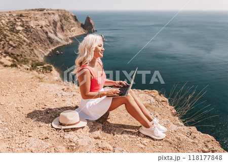 A woman is sitting on a rocky beach with a laptop open in front of her. She is wearing a pink top and white skirt. Concept of relaxation and leisure. A woman is sitting on a rocky beach with a laptop open in front of her. She is wearing a pink top and white skirt. Concept of relaxation and leisure. 118177848