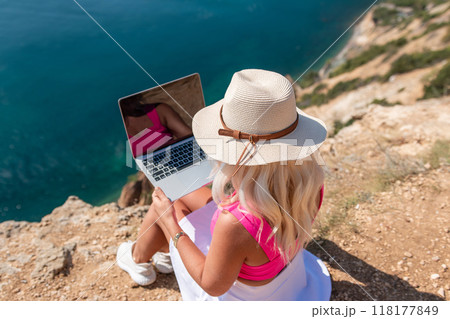 A woman is sitting on a rocky beach with a laptop in front of her. She is wearing a pink top and a straw hat. Concept of relaxation and leisure. A woman is sitting on a rocky beach with a laptop in front of her. She is wearing a pink top and a straw hat. Concept of relaxation and leisure. 118177849