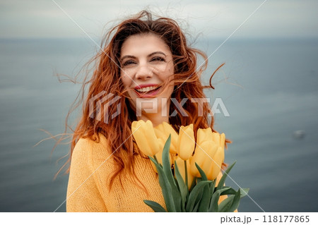 Portrait of a happy woman with hair flying in the wind against the backdrop of mountains and sea. Holding a bouquet of yellow tulips in her hands, wearing a yellow sweater 118177865
