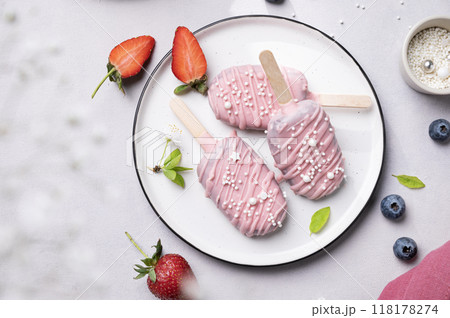 Cake pops ice cream on a stick in pink glaze on a plate with berries on a light background.  118178274