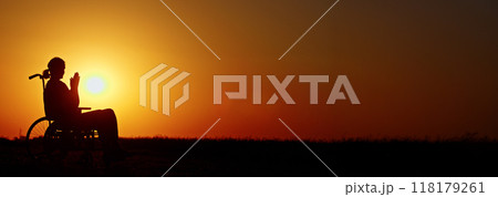Silhouette of a person in a wheelchair at sunset with hands clasped, set against a vivid orange sky. 118179261