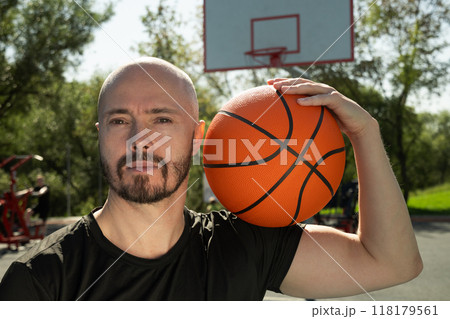 Portrait of sporty man holding basketball orange ball on shoulder and looking at camera on sports court. Basketball coach. Close up. Outdoors. Portrait of sporty man holding basketball orange ball on shoulder and looking at camera on sports court. Basketball coach. Close up. Outdoors. 118179561