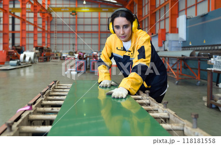 Young female hispanic labor dressed in polyester jacket uniform and ear muff working in metal sheet manufacture. Various kind of industrial machine are in the working area. Young female hispanic labor dressed in polyester jacket uniform and ear muff working in metal sheet manufacture. Various kind of industrial machine are in the working area. 118181558