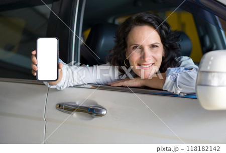 Caucasian woman is sitting in the driver seat of a car with the window down. She is smiling and holding a smartphone with a blank screen towards the camera. 118182142