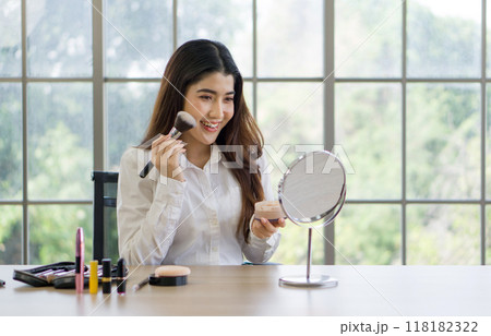 A woman is happily applying makeup while looking in a mirror at a table with various beauty products. A woman is happily applying makeup while looking in a mirror at a table with various beauty products. 118182322