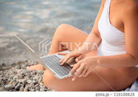 A woman is sitting on a beach with a laptop open in front of her. She is typing on the keyboard and she is focused on her work. A woman is sitting on a beach with a laptop open in front of her. She is typing on the keyboard and she is focused on her work. 118188628