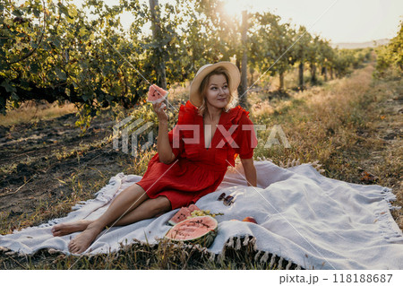 A woman in a red dress is sitting on a blanket in a field with a watermelon in her hand. A woman in a red dress is sitting on a blanket in a field with a watermelon in her hand. 118188687