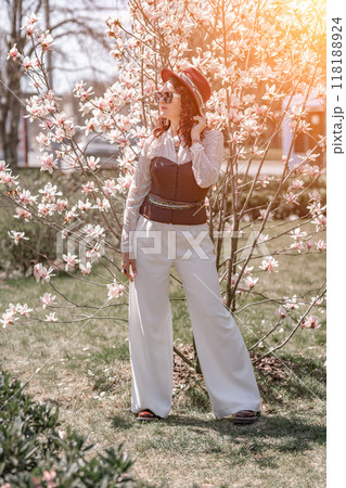 Magnolia park woman. Stylish woman in a hat stands near the magnolia bush in the park. Dressed in white corset pants and posing for the camera. Magnolia park woman. Stylish woman in a hat stands near the magnolia bush in the park. Dressed in white corset pants and posing for the camera. 118188924