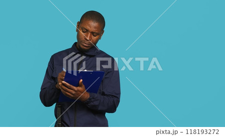 Security guard holding clipboard, taking notes, receiving orders during protection job shift. Portrait of bodyguard using pen to write informations received, studio background, camera A Security guard holding clipboard, taking notes, receiving orders during protection job shift. Portrait of bodyguard using pen to write informations received, studio background, camera A 118193272