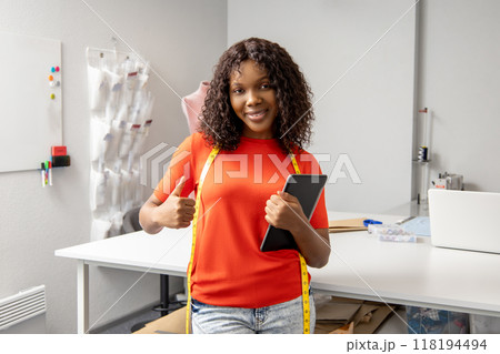 Woman tailor holding tablet with measuring tape around neck in well-equipped atelier 118194494