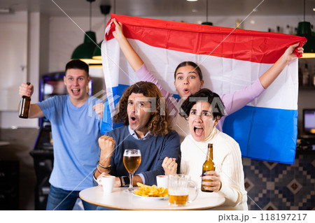 Company of excited young adult sports fans waving Dutch flag and supporting national team with beer in pub Company of excited young adult sports fans waving Dutch flag and supporting national team with beer in pub 118197217
