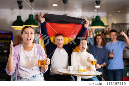 Company of emotional young adult fans supporting German team with state flag while drinking beer in sports bar Company of emotional young adult fans supporting German team with state flag while drinking beer in sports bar 118197310