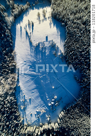 Aerial view top-down reveals snow-covered landscape with small cluster of buildings surrounded by dense pine forests. Long shadows of trees and structures create intricate patterns on pristine snow. 118201718