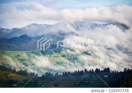 Picturesque scene of mountain valley blanketed in mist, with patches of green pastures and scattered houses. Clouds drift gently over forested hills, creating serene and tranquil atmosphere. 118201728