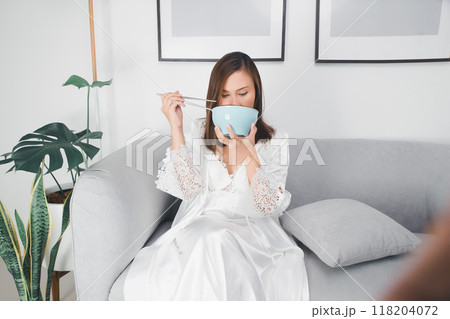 At midnight, an Asian woman, hungry and tired, sits on her couch eating food from a bowl. 118204072