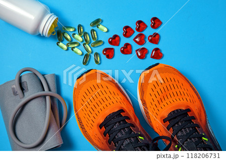 Tonometer for measuring blood pressure, sports orange sneakers, yellow vitamin D capsules and red glass hearts scattered on a blue background. Vitamin supplements and exercise for heart health 118206731