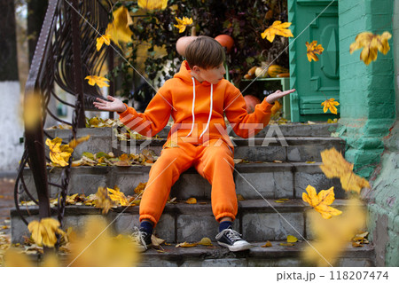 A boy in orange clothes sits on the steps and looks at the yellow flying autumn leaves. Fall has come. 118207474