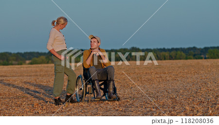 A woman talks to a young man in a wheelchair in an open field at sunset. A woman talks to a young man in a wheelchair in an open field at sunset. 118208036