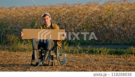 A young man in a wheelchair holds a blank cardboard sign in front of a cornfield. 118208038