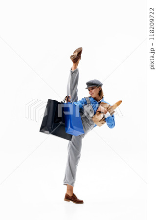 Gracefully lifting leg high, woman in dynamic ballet pose while balancing shopping bags and loaf of bread against white background. Gracefully lifting leg high, woman in dynamic ballet pose while balancing shopping bags and loaf of bread against white background. 118209722