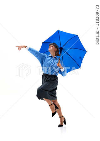 Stylish woman, dressed retro outfit, leaps with blue umbrella, gracefully pointing forward in playful pose against white studio background. Stylish woman, dressed retro outfit, leaps with blue umbrella, gracefully pointing forward in playful pose against white studio background. 118209773