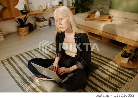 Gen Z female writer with albinism sitting on floor in cozy living room and working on laptop in morning 118210052