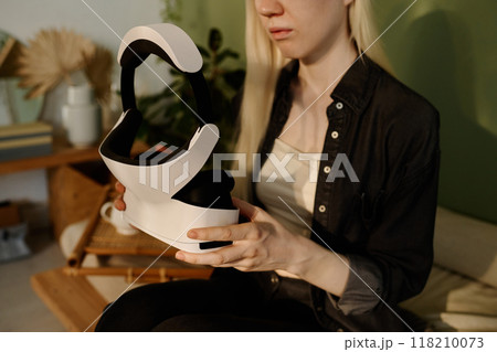 Medium section shot of young woman with long white hair holding modern VR headset in her hands while sitting on couch at home 118210073