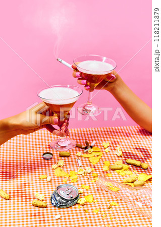 Two hands hold cocktail glasses with cigarette between fingers, surrounded by scattered snacks on gingham tablecloth against pink background. Two hands hold cocktail glasses with cigarette between fingers, surrounded by scattered snacks on gingham tablecloth against pink background. 118211879
