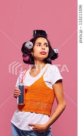 Woman looks away with playful expression, her hairstyle made of cans and drink in hand against pink background. Playful styling. Woman looks away with playful expression, her hairstyle made of cans and drink in hand against pink background. Playful styling. 118211896