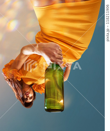 Young beautiful woman in sunny yellow dress holds large green bottle of sparkle wine posing against clear blue sky. Creative photo. 118211906