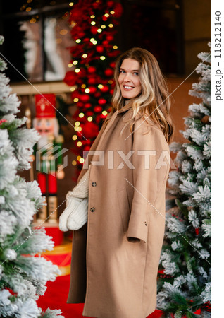 portrait of a happy woman in a coat and white mittens posing against the backdrop of Christmas decorations, the mood of the approaching holiday 118212140