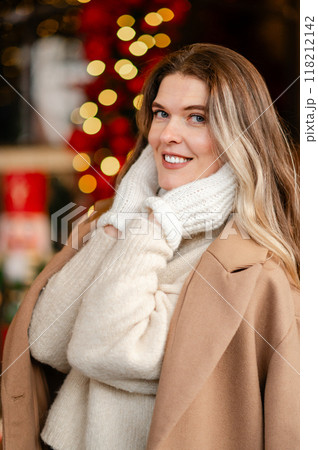 portrait of a happy woman in a coat and white mittens posing against the backdrop of Christmas decorations, the mood of the approaching holiday portrait of a happy woman in a coat and white mittens posing against the backdrop of Christmas decorations, the mood of the approaching holiday 118212142