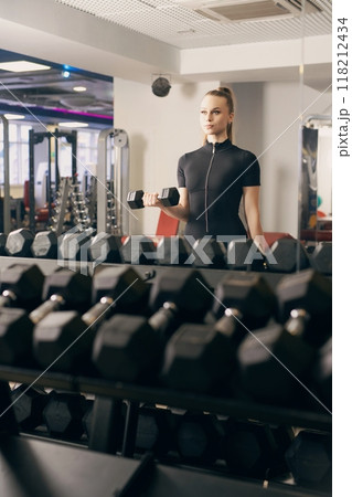 Woman lifting dumbbell in modern gym with reflected weights during workout 118212434