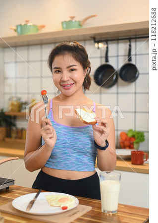 Pretty young asian woman in sportswear eating toasted bread for breakfast in kitchen 118212688