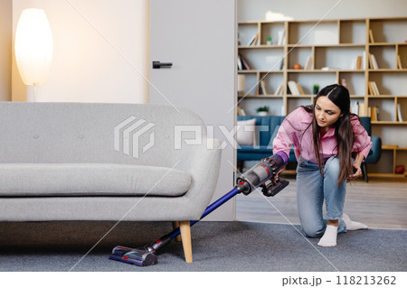 Young woman uses cordless vacuum cleaner to clean home carpet. Modern easy cleaning 118213262