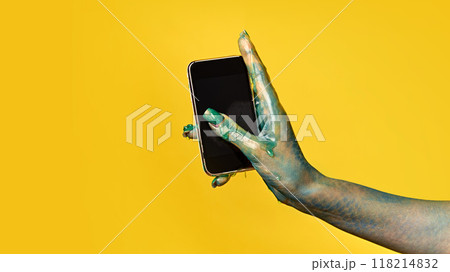 Hand covered in green slime holding black smartphone with blank screen against yellow background. Halloween-themed image for social media and marketing materials. 118214832