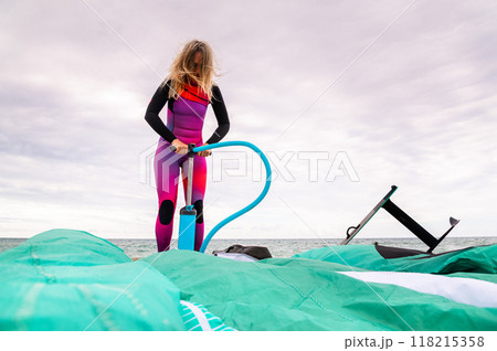Young sportswoman in a wetsuit pumps her kite on the beach in cloudy weather 118215358